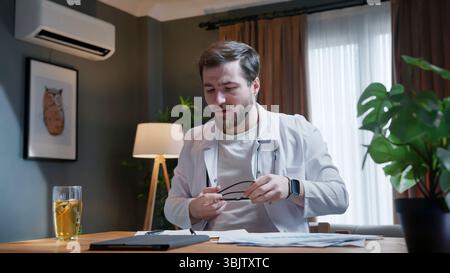 Junger männlicher Arzt, der eine Brille hält und am Schreibtisch sitzt, sich auf eine Fernkonsultation vorbereitet oder Patientendokumente überprüft. Stockfoto