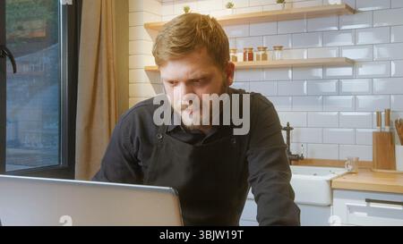 Bärtiger Mann mit schwarzer Schürze schaut aufmerksam auf einen Laptop-Bildschirm und schlägt Arbeit, Forschung oder Planung in einer Küche vor. Stockfoto