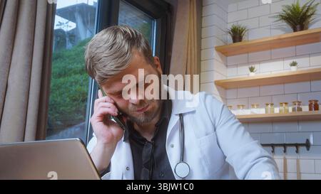 Ein bärtiger Arzt im weißen Mantel spricht am Telefon mit einem ernsten Ausdruck, während er an einem Laptop in der Nähe eines Fensters im Büro arbeitet. Stockfoto