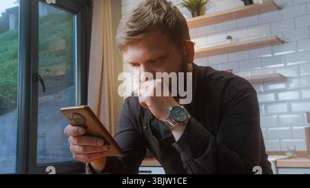 Ein bärtiger Mann in schwarzem Hemd sitzt am Fenster, schaut mit einem besorgten Ausdruck auf das Smartphone und liest möglicherweise schlechte Nachrichten oder finanzielle Updates. Stockfoto