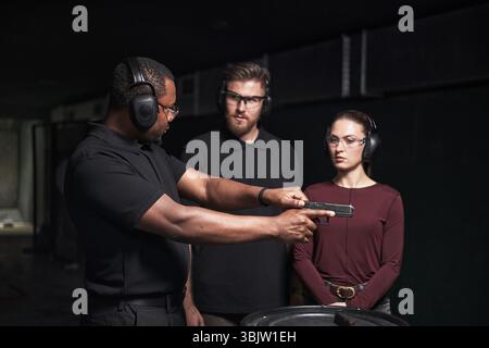 Afroamerikanischer Mann, der kaukasische junge Erwachsene Männer und Frauen im Schießklub unterweist, alle tragen Gehörschützer und Schutzbrille, wobei der Schwerpunkt auf der Handhabung von Handfeuerwaffen liegt Stockfoto