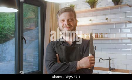 Ein bärtiger Mann, der eine schwarze Schürze trägt, lächelt selbstbewusst, während er ein Küchenmesser hält und in einer hellen, modernen Küche steht. Stockfoto