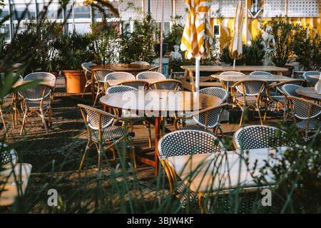 Gefaltete und geschlossene große Sonnenschirme, Tische und Stühle im Straßenrestaurant oder Café in Europa. Sonniger Sommermorgen vor der Öffnungszeit, keine Leute Stockfoto