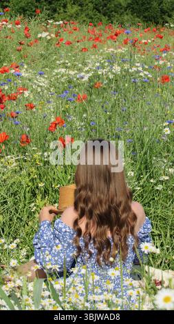 Eine junge Frau sitzt friedlich auf einer farbenfrohen Wildblumenwiese, umgeben von blühenden Mohnblumen und Gänseblümchen. Sie hält einen Strohhut und saugt die Wärme auf Stockfoto