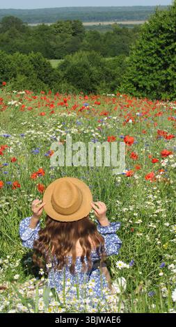 Eine Frau sitzt friedlich in einem farbenfrohen Wildblumenfeld und passt ihren Strohhut an, während sie von blühenden roten Mohnblumen und blauen Blumen umgeben ist. Stockfoto