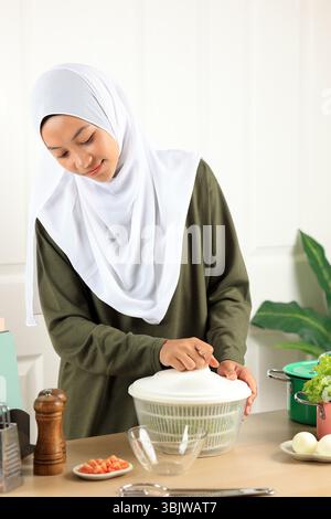 Frau in Hijab entwässert Salat aus dem Wasser in einer Salatschleuder, und macht Salat in der Küche nach und nach Stockfoto