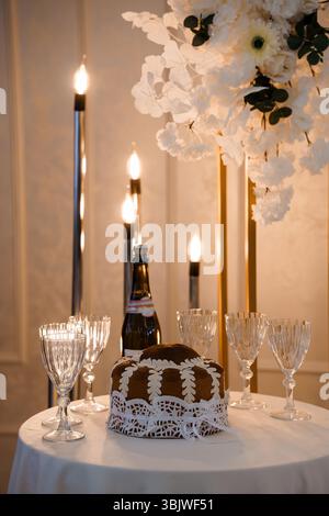 Ein wunderschön gestalteter Hochzeitstisch verfügt über einen zweistufigen Kuchen, der mit Spitze, Sektflasche, Kristallgläsern und flackernden Kerzen verziert ist Stockfoto