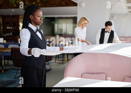 Junge Erwachsene Schwarze Frau Setting Table mit Kollegen im Restaurant Interior Stockfoto