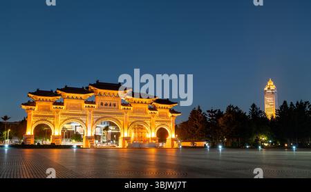 Weite Dämmerung Blick auf den Liberty Square Arch, der von goldenen Flutlichtern erstrahlt, und seine kunstvollen traditionellen Dachsilhouetten spiegeln sich von einem entfernten Turm hinter dem geräumigen plaza wider Stockfoto