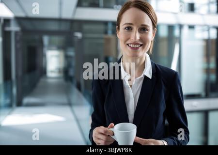 Rothaarige Executive mit marineblauem Blazer, weißem Kaffeetasse und lächelndem Lächeln im Glaskorridor Stockfoto