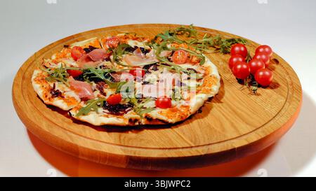 Köstliche Pizza mit Schinken und Kirschtomaten auf einem Holzbrett. Eine Nahaufnahme einer frisch gebackenen Pizza mit Schinkenscheiben und Kirschfleisch Stockfoto