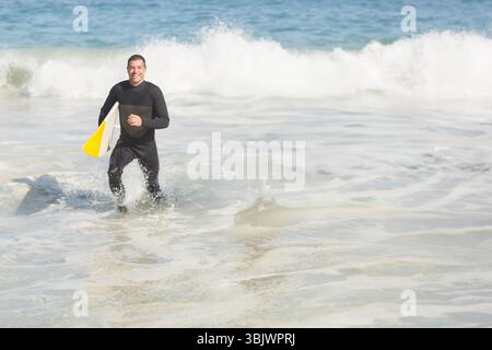 Mann mittleren Alters in schwarzem Neoprenanzug, der am Strand aus dem Meer auftaucht und weißes und gelbes Surfbrett trägt Stockfoto