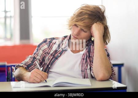 Männlicher Schüler schreibt in Notizbuch mit Stift am Schreibtisch im Klassenzimmer unter hellem Fensterlicht Stockfoto