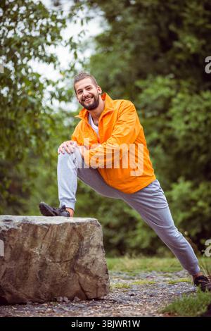 Mann mit leuchtend orangefarbenem Windbreaker und Sneakers, der das Bein auf Felsen in der Waldlichtung spannt Stockfoto