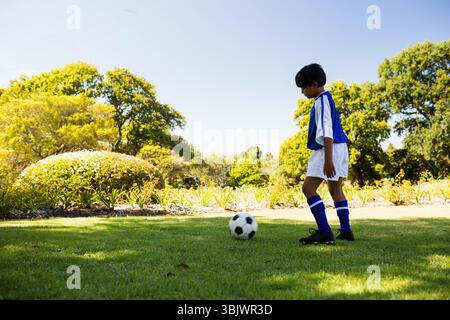 Kind in blau-weißer Uniform, das auf Gras in der Nähe von Sträuchern steht und sich auf schwarz-weißen Ball konzentriert Stockfoto