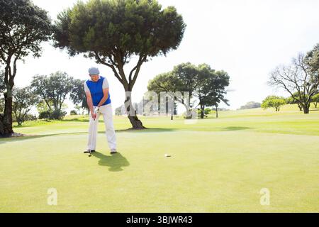 Mann, der Golfputt auf sonnendurchflutetem Grün mit Ball in der Nähe des Lochs anstellt, mit klarem Himmel über dem Kopf Stockfoto