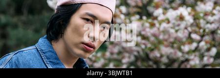 Ein junger asiatischer Mann zeigt sein trendiges Denim-Outfit unter blühenden Blumen im Park, Banner Stockfoto