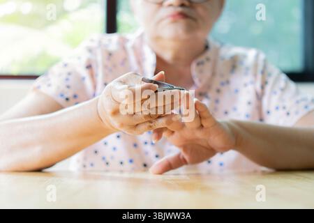 Seitenaufnahme einer älteren asiatischen Frau, die zu Hause sitzt und sich die Fingernägel schneidet. Selektiver Fokus. Stockfoto