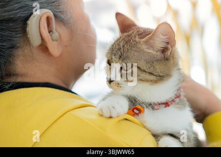 Fröhliche Seniorin mit Hörgerät am Fenster, während sie eine Katze auf der Schulter hält. Freundschaft mit Cat-Konzept. Stockfoto