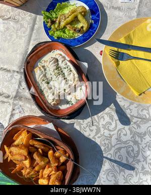 Köstliches traditionelles Balkan-Mittagessen mit gegrillter Paprika, cremigem Hähnchenfilet und knusprigen Kartoffeln, serviert auf einem rustikalen Tisch bei natürlichem Sonnenlicht. Stockfoto