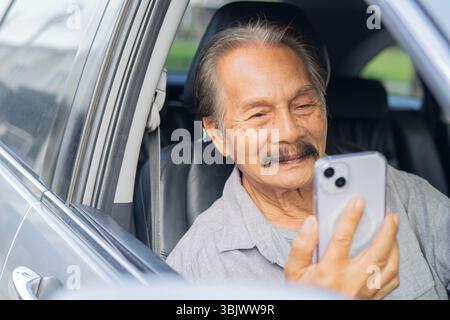 Lächelnder Seniorenmann mit Bart, der Auto fährt und ein Smartphone hält an einem sonnigen Tag im Freien. Reifer Fahrer mit Glückskonzept Stockfoto