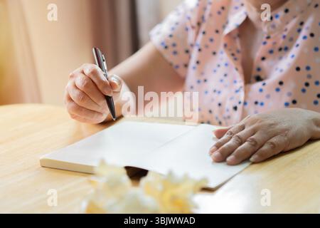 Eine alte Frau schreibt mit einem Stift auf ein Buch. Schuss von einer älteren Frau, die zu Hause in ein Tagebuch schreibt. Selektiver Fokus. Stockfoto