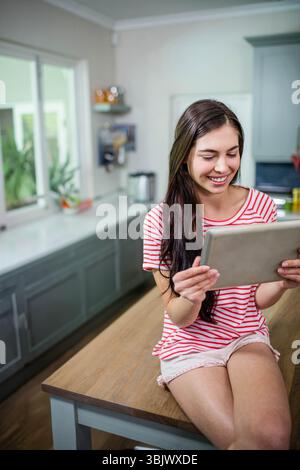 Frau, die auf hölzernen Kücheninsel sitzt, Tablette in der Küche in der Nähe von Topfpflanzen hält, Kopierraum Stockfoto