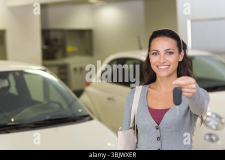 Frau mit Autoschlüssel und beigefarbener Tasche im Showroom mit weißen Autos, Kopierraum Stockfoto