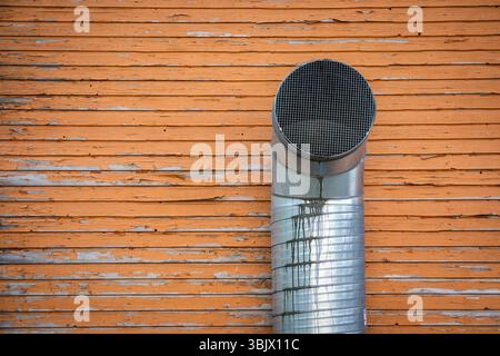 Industrielles Lüftungsrohr auf verwitterter Holzwand mit abziehender orangefarbener Farbe. Stockfoto