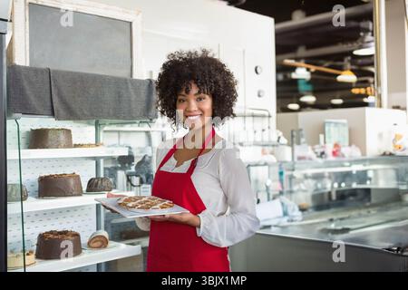 Afroamerikanerin Bäckerin in der Bäckerei mit Schürze, die Gebäck auf Tablett hinter Glaskasten präsentiert Stockfoto