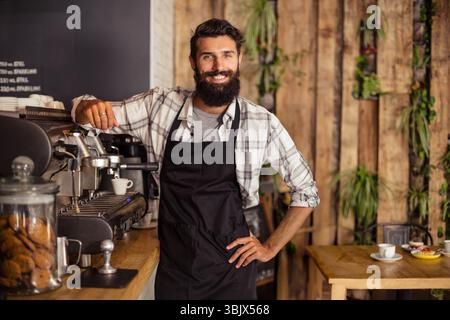 Männlicher Barista in schwarzer Schürze, der sich im Café auf eine Holztheke stützt und Espresso mit Keksdose zubereitet Stockfoto