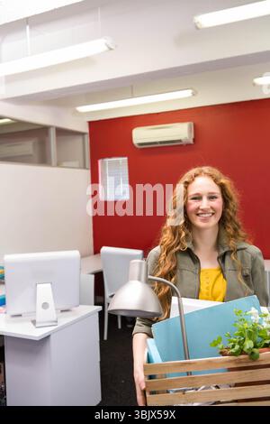 Weibliche Büroangestellte hält Holzkiste mit Lampe, Pflanze und Ordnern im offenen Büro, Kopierraum Stockfoto