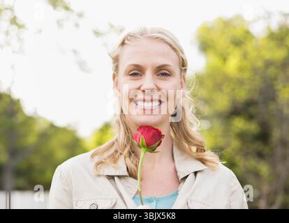 Mittelerwachsene Frau, die eine rote Rose hält und draußen im Garten mit grünem Laub lächelt Stockfoto
