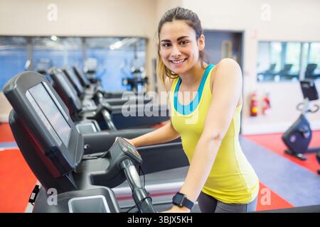 Hispanische Frau im Fitnessstudio mit Laufband, lächelnd, mit Konsole, Spiegelwand mit Cardio-Fahrrädern Stockfoto