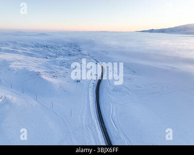 Ein Blick aus der Vogelperspektive auf eine kurvenreiche Straße inmitten einer schneebedeckten Landschaft mit tief liegendem Nebel oder Wolken in der Ferne Stockfoto