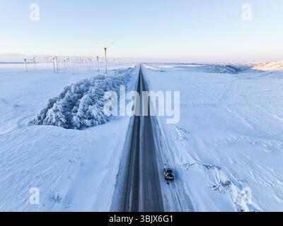 Ein Blick auf ein Auto auf einer langen, geraden Straße durch eine schneebedeckte Landschaft mit einem Windpark im Hintergrund Stockfoto