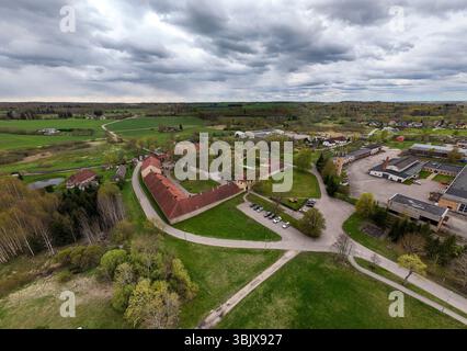 Slokenbeka, Lettland - 26. April 2025: Aus der Vogelperspektive ist eine malerische Stadt umgeben von üppigen grünen Feldern unter grauen Wolken. Stockfoto