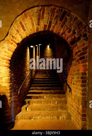 Eine Treppe nach oben ist von einem Backsteinbogen eingerahmt, wobei die Stufen und Mauern aus Backstein bestehen. Die Szene wird durch Deckenleuchten beleuchtet, die gegossen werden Stockfoto