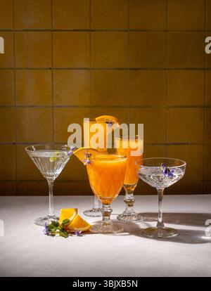 Leuchtend orangefarbene Cocktails, dekoriert mit essbaren Blumen und orangen Scheiben, stehen auf einer weißen Tischdecke vor einer gelb gefliesten Wand Stockfoto