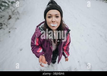 Frau genießt Winterspaß im Freien, hält und probiert Schnee, warm gekleidet in einer lila Jacke und Mütze, umgeben von einer verschneiten Landschaft. Stockfoto