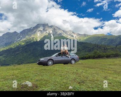 Junge Paare sitzen auf dem Dach ihres Autos und genießen den atemberaubenden Panoramablick auf eine majestätische Bergkette während eines unvergesslichen Roadtrips Stockfoto