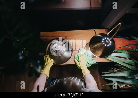 Blick von oben auf eine Person, die Holzmöbel mit einem gelben Handschuh und einem grünen Tuch unter warmem Lampenlicht reinigt Stockfoto