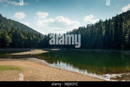 Synevyr See in Karpaten. Der See ist von hohen Kiefern umgeben. Reflexion der Wald- und Hügelkette im Wasser. Teilweise bewölkter Himmel. Stockfoto