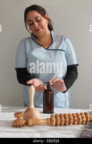Hispanische Massagetherapeutin in professioneller Uniform-Putting-Lotion, Vorbereitung und Organisation von Holzmassagewerkzeugen, auf einem Massagetisch in einem Kalk Stockfoto