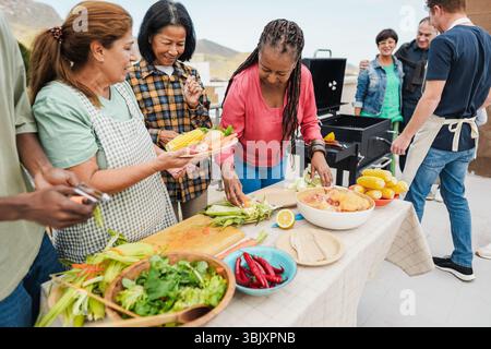 Glückliche Frauen, die Speisen auf dem Dach zu Hause zubereiten - Seniorenfreunde, die Spaß beim gemeinsamen Kochen am Wochenende haben - Sommer Stockfoto