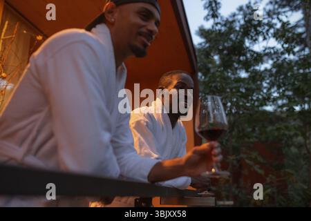 Afroamerikanisches schwules männliches Paar genießt ein romantisches Resort-Wochenende und einen schönen Abend, spricht und trinkt Wein auf einer Terrasse, ein Niedrigwinkelfoto. Stockfoto