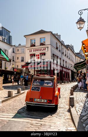 Ein orangefarbener Citroën 2CV fährt durch Montmartre mit dem berühmten Restaurant „le Consulat“ im Hintergrund. Ein Reiseführer mit Touristen in einem Citroën Stockfoto