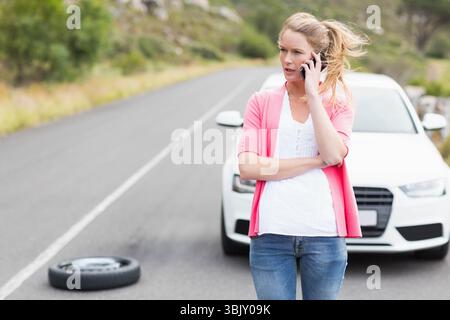 Frau, die auf einer asphaltierten Landstraße neben weißer Limousine steht, Smartphone und Ersatzreifen hält, Kopierraum Stockfoto