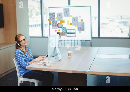 Frau mit Headset, die am Monitor am Holztisch im Büro mit Kaffeetassen und Kopierraum arbeitet Stockfoto