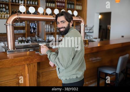Bärtiger Mann in grüner Jacke, der an der Bar steht und einen Bierbecher in der Nähe von Kupferhähnen hält Stockfoto